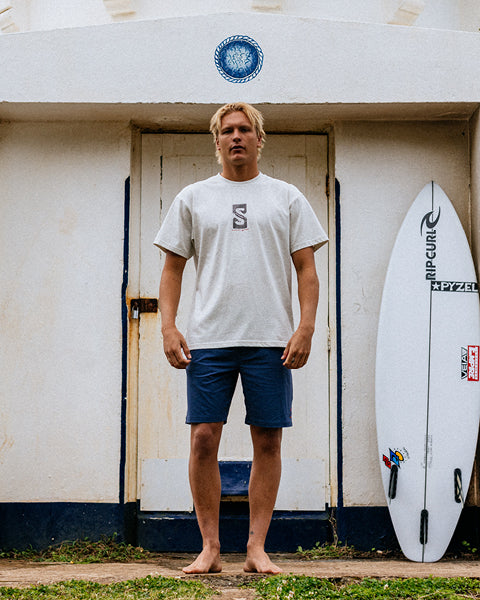 Man standing next to a surfboard with a shed and surfboard in the background