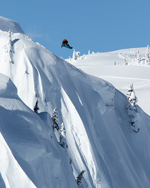Person snowboarding down a steep, snow-covered mountain with a clear blue sky.