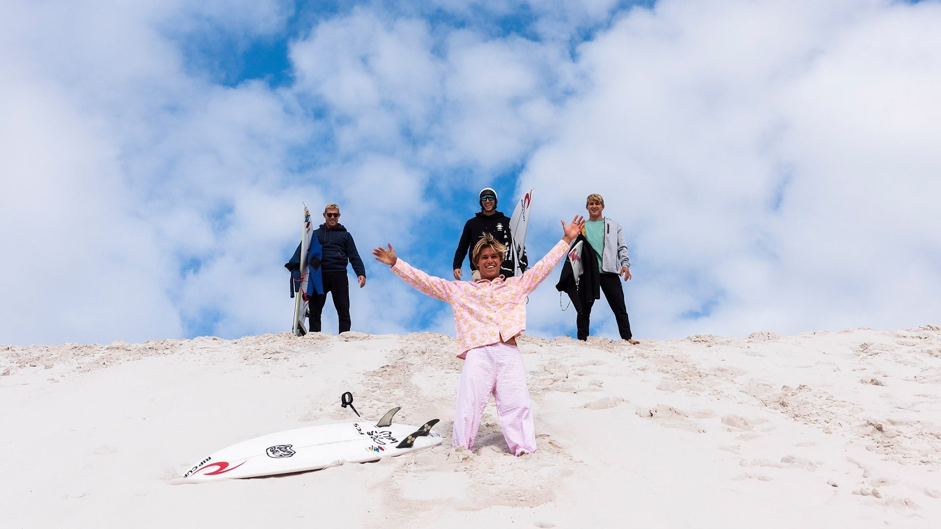Group of people with snowboards on a snowy slope under a blue sky with clouds.