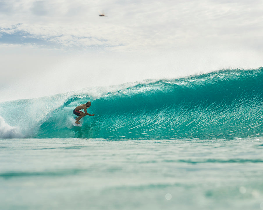 Person surfing a large wave in clear blue water with a cloudy sky.