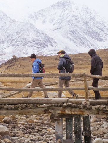 Three people walking on a wooden bridge with snow-capped mountains in the background