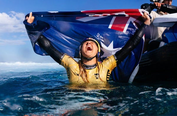 Person in a yellow wetsuit holding an Australian flag above their head in the ocean.
