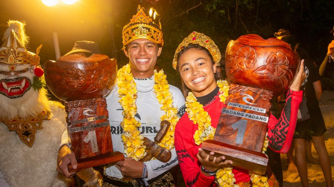 Two individuals holding large wooden bowls with decorative crowns and leis, surrounded by a festive atmosphere.
